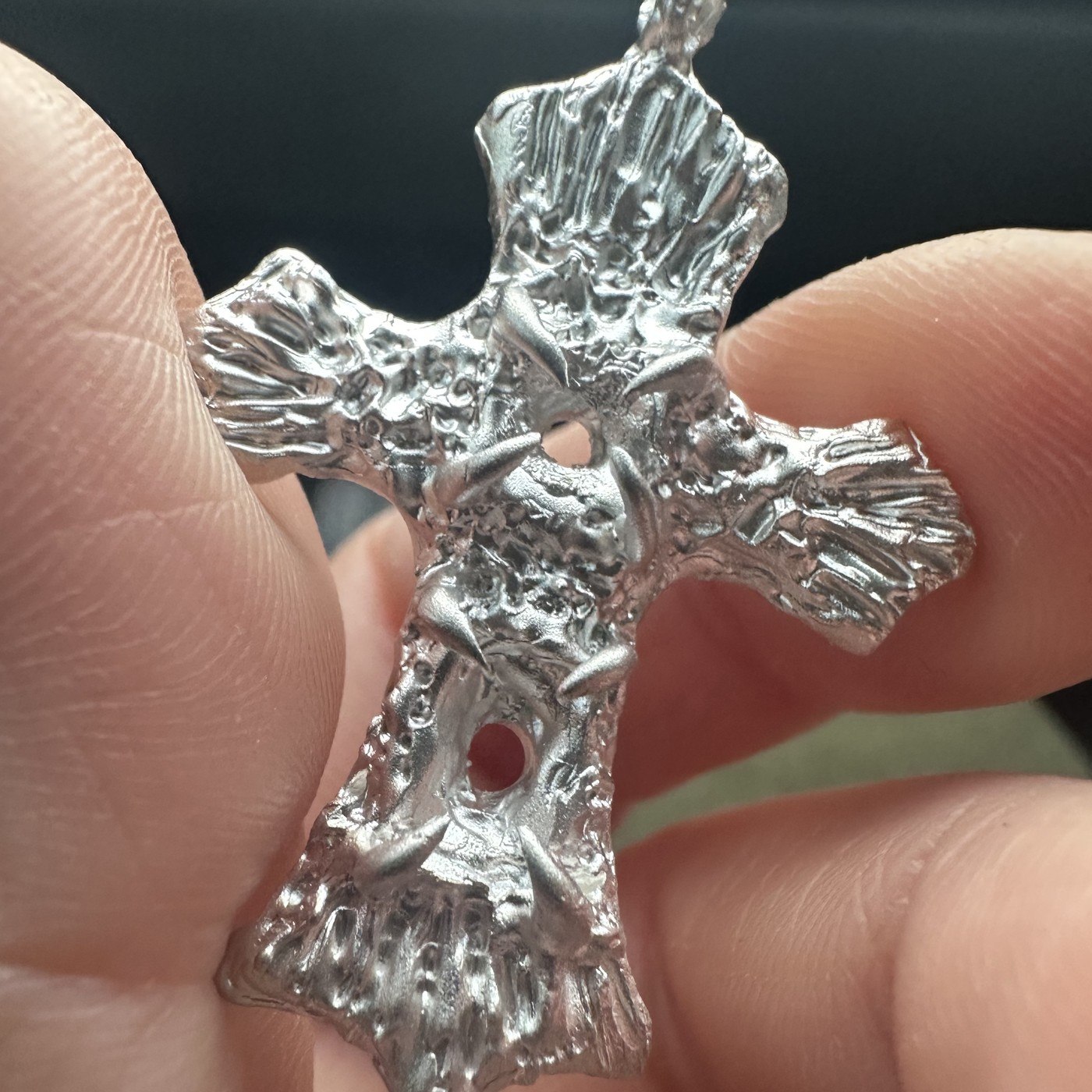 A raw silver cross pendant, freshly cast and not yet polished, held between the fingers